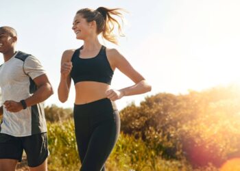 A man and a woman jogging outdoors on a sunny day, with trees and vegetation in the background.