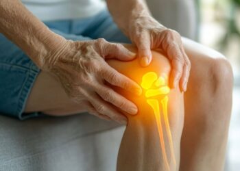 Elderly person holding a glowing knee joint, indicating pain or discomfort while sitting on a couch.
