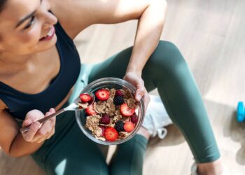 Person sitting on the floor, holding a bowl of granola, strawberries, and blackberries, next to a blue dumbbell.
