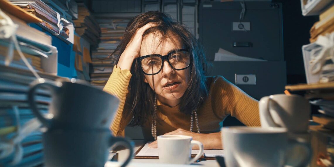 A person wearing glasses looks stressed, surrounded by stacks of papers and several coffee cups on a desk.