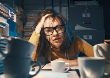 A person wearing glasses looks stressed, surrounded by stacks of papers and several coffee cups on a desk.