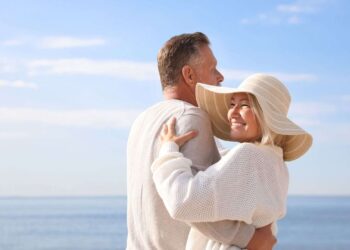 A middle-aged couple stands embracing by the seaside on a sunny day; the woman is smiling and wearing a wide-brimmed hat.