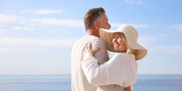 A middle-aged couple stands embracing by the seaside on a sunny day; the woman is smiling and wearing a wide-brimmed hat.