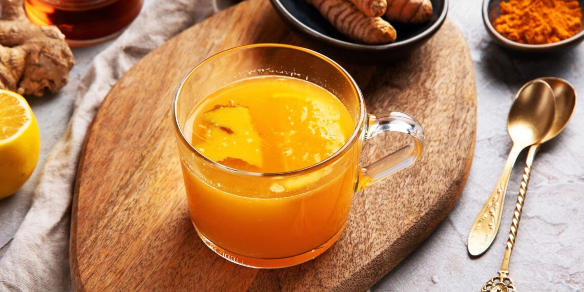 A glass mug of orange-yellow turmeric tea sits on a wooden board, surrounded by fresh turmeric, ginger, a lemon half, turmeric powder, and a gold spoon.