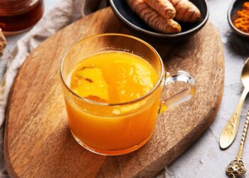 A glass mug of orange-yellow turmeric tea sits on a wooden board, surrounded by fresh turmeric, ginger, a lemon half, turmeric powder, and a gold spoon.