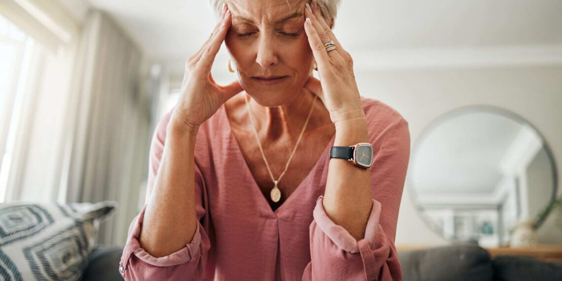 Older woman sitting on a sofa with eyes closed, touching her temples with both hands, appearing to have a headache or stress.