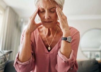 Older woman sitting on a sofa with eyes closed, touching her temples with both hands, appearing to have a headache or stress.