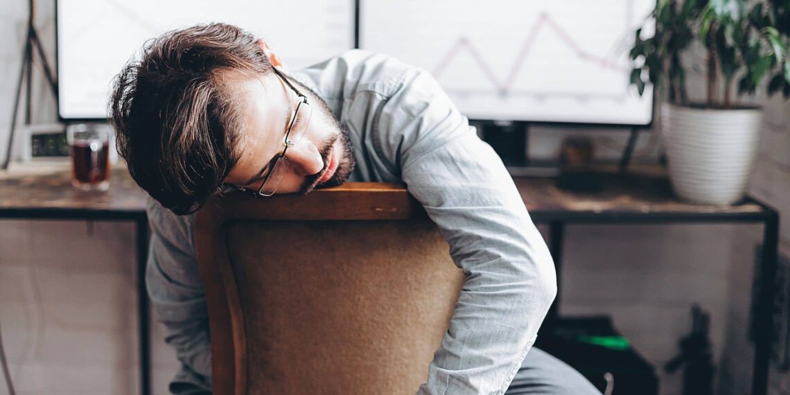 A man with glasses sits slumped over a chair in front of a desk with monitors displaying graphs and a potted plant nearby.