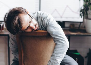 A man with glasses sits slumped over a chair in front of a desk with monitors displaying graphs and a potted plant nearby.