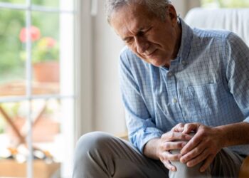 An older man sitting indoors holds his knee with both hands and appears to be in pain.
