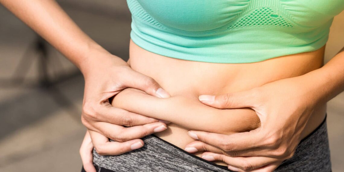 A person wearing a green sports bra and gray pants pinching the skin and fat on their bare abdomen with both hands.