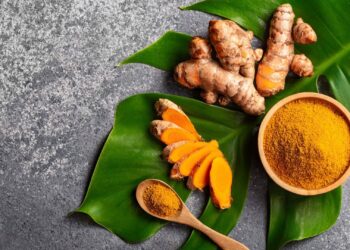Whole turmeric roots, sliced turmeric, a bowl of turmeric powder, and a wooden spoon with powder arranged on green leaves over a gray surface.
