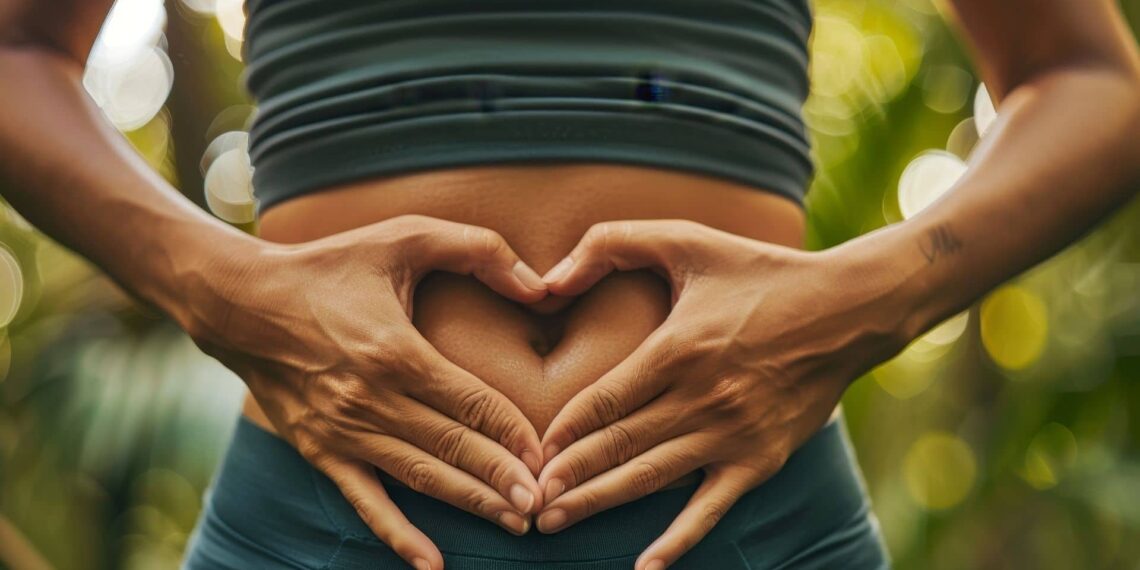 A person forms a heart shape with their hands over their bare abdomen, standing outdoors with greenery in the background.