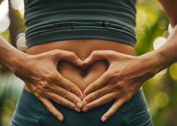 A person forms a heart shape with their hands over their bare abdomen, standing outdoors with greenery in the background.