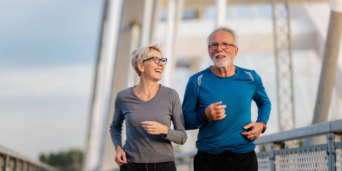 An older woman and man are jogging together outdoors on a bridge, both dressed in athletic wear and appearing cheerful.