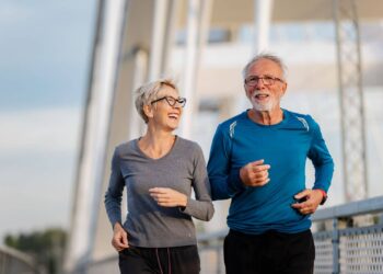 An older woman and man are jogging together outdoors on a bridge, both dressed in athletic wear and appearing cheerful.