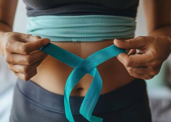 Person tying a blue measuring tape around their bare midsection while wearing athletic clothing.