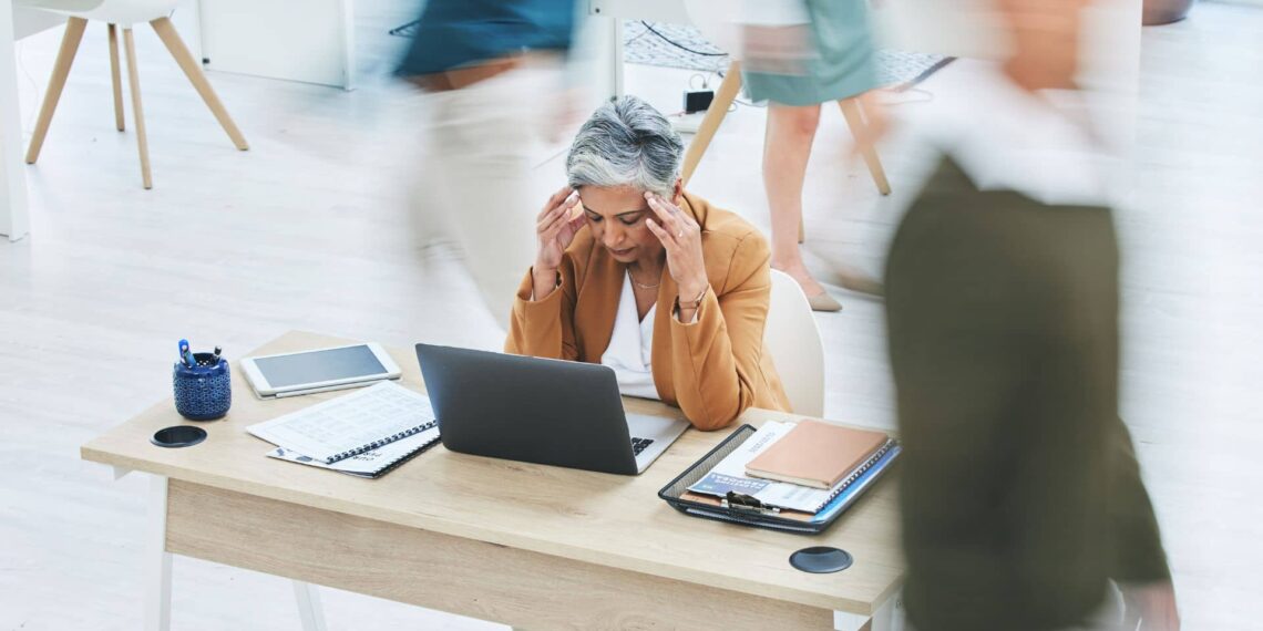 A woman sits at a desk with her hands on her temples, appearing stressed, surrounded by office supplies and blurred people walking past in a busy office setting.