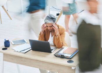 A woman sits at a desk with her hands on her temples, appearing stressed, surrounded by office supplies and blurred people walking past in a busy office setting.