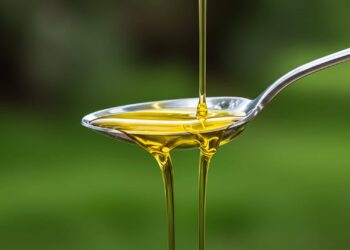 A stream of yellow oil is being poured into a shiny metal spoon, with a green blurred background.