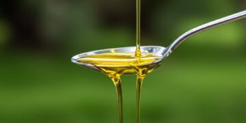 A stream of yellow oil is being poured into a shiny metal spoon, with a green blurred background.