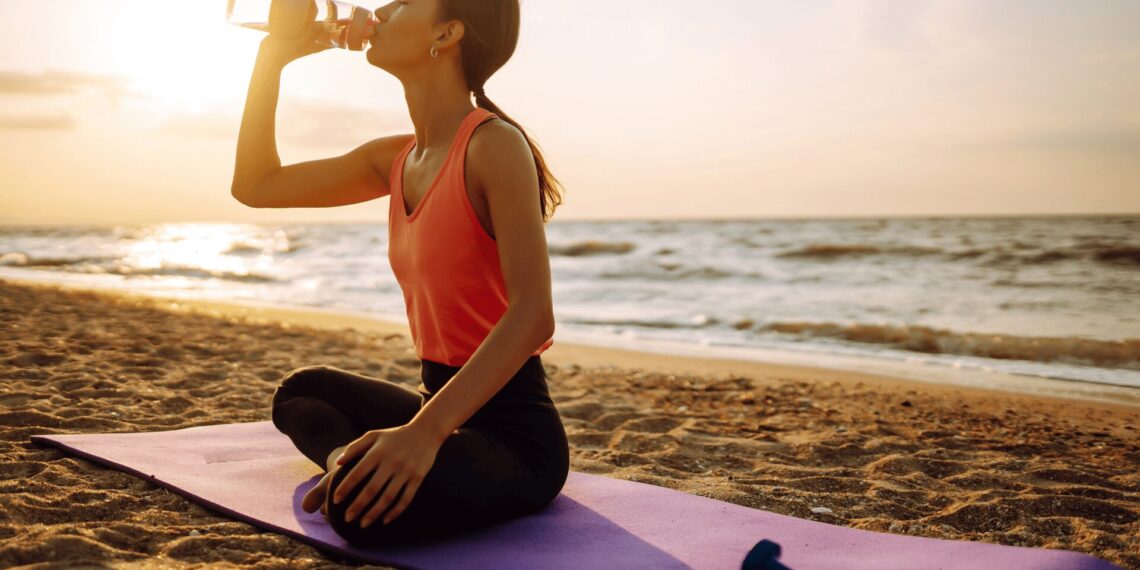 A person sits on a yoga mat on the beach at sunset, drinking from a water bottle, with a pair of dumbbells nearby.