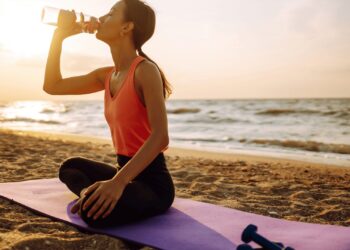 A person sits on a yoga mat on the beach at sunset, drinking from a water bottle, with a pair of dumbbells nearby.