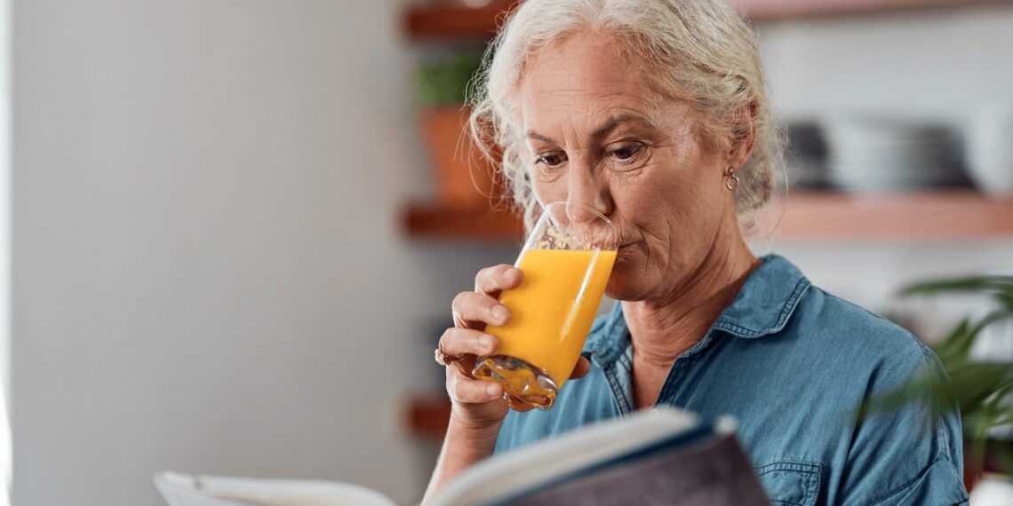 Older woman with gray hair wearing a blue shirt drinks orange juice while reading a book indoors.