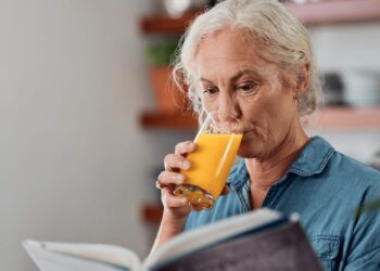 Older woman with gray hair wearing a blue shirt drinks orange juice while reading a book indoors.