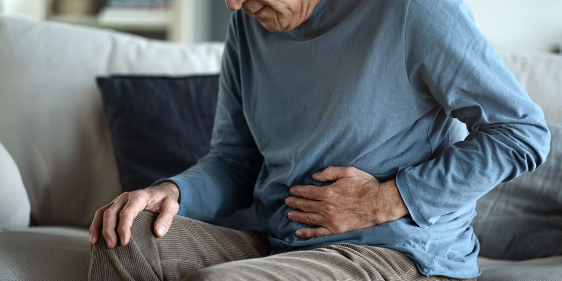 An older person sitting on a couch holds their abdomen with one hand, appearing to experience stomach pain or discomfort.