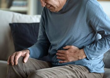 An older person sitting on a couch holds their abdomen with one hand, appearing to experience stomach pain or discomfort.
