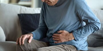 An older person sitting on a couch holds their abdomen with one hand, appearing to experience stomach pain or discomfort.