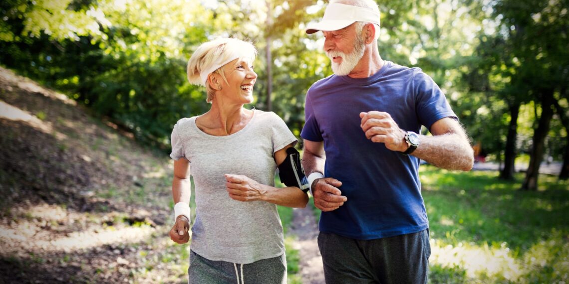 Older man and woman jogging outdoors on a sunny day, wearing casual athletic clothes and smiling at each other. Trees and greenery are visible in the background.
