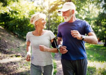 Older man and woman jogging outdoors on a sunny day, wearing casual athletic clothes and smiling at each other. Trees and greenery are visible in the background.