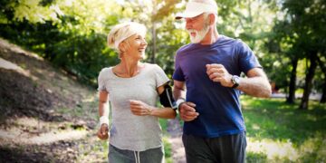 Older man and woman jogging outdoors on a sunny day, wearing casual athletic clothes and smiling at each other. Trees and greenery are visible in the background.