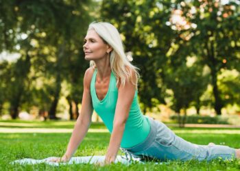 A woman in athletic wear practices yoga outdoors on a grassy field, performing an upward facing dog pose with trees in the background.